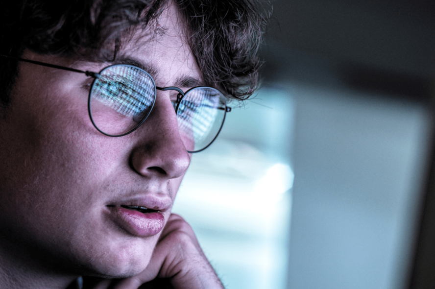 Close up of a data science student looking at a computer screen; a screen full of data is reflected in his eyeglasses.