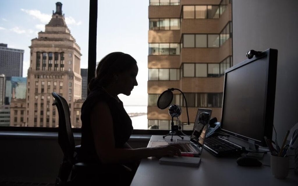A profile of a person sitting in front of a window, working on her computer.