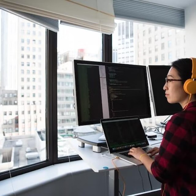 A student wearing headphones sits in front of multiple computer screen monitors.