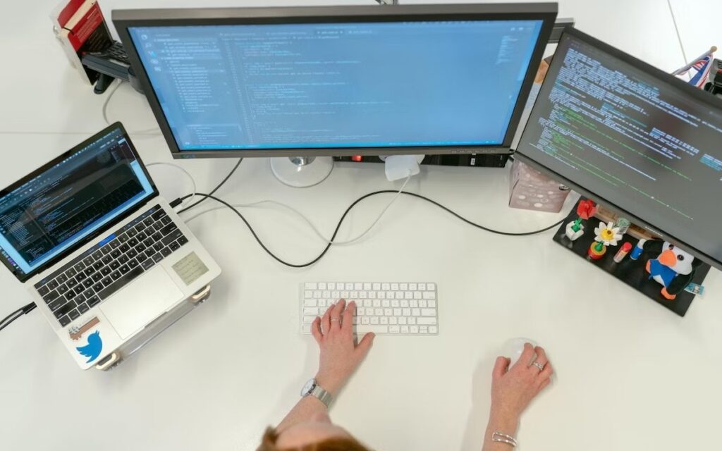 A person types on a white keyboard while surrounded by three computer monitors.