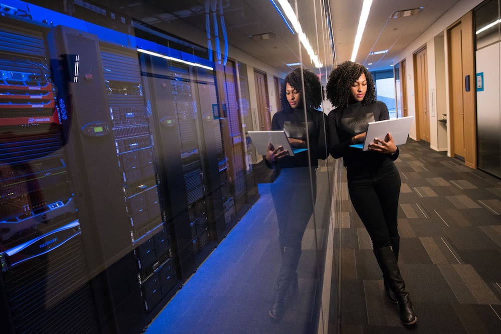 A Black woman holding and looking at a small laptop leans against a glass wall encasing a bank of computer servers.