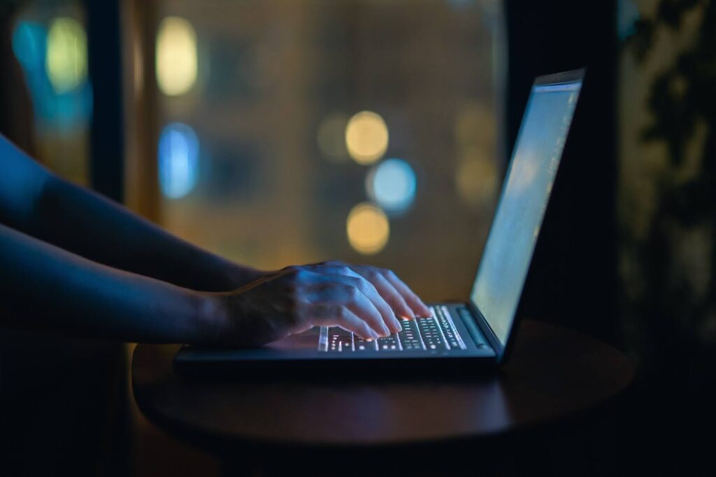 Close up of hands typing at a laptop computer in a darkened room.