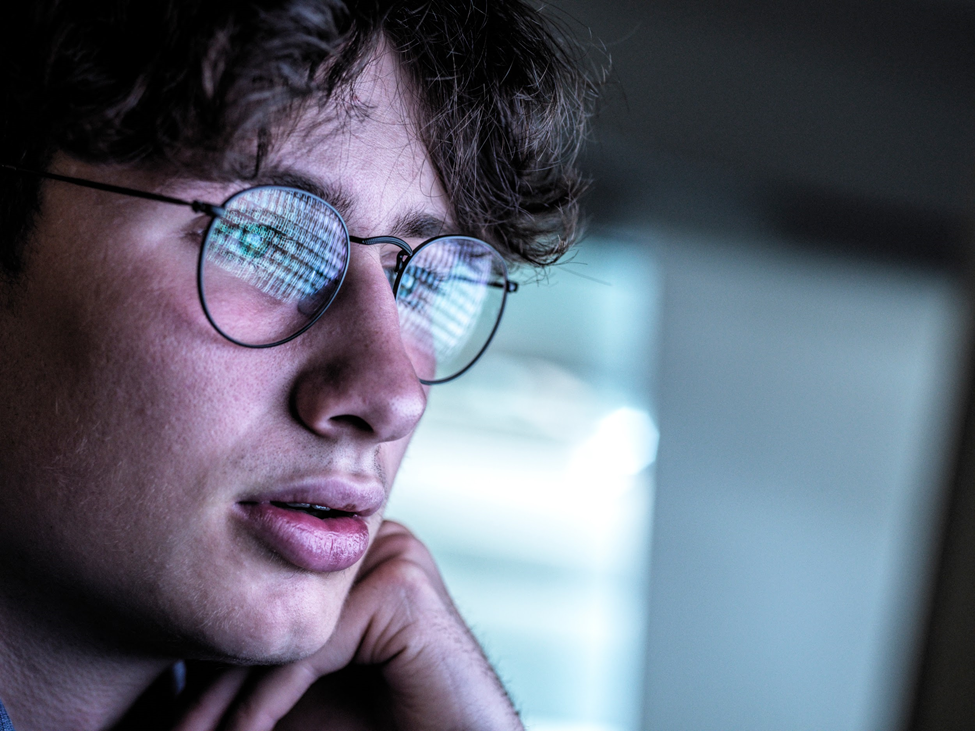 Close up of a data science student looking at a computer screen; a screen full of data is reflected in his eyeglasses.