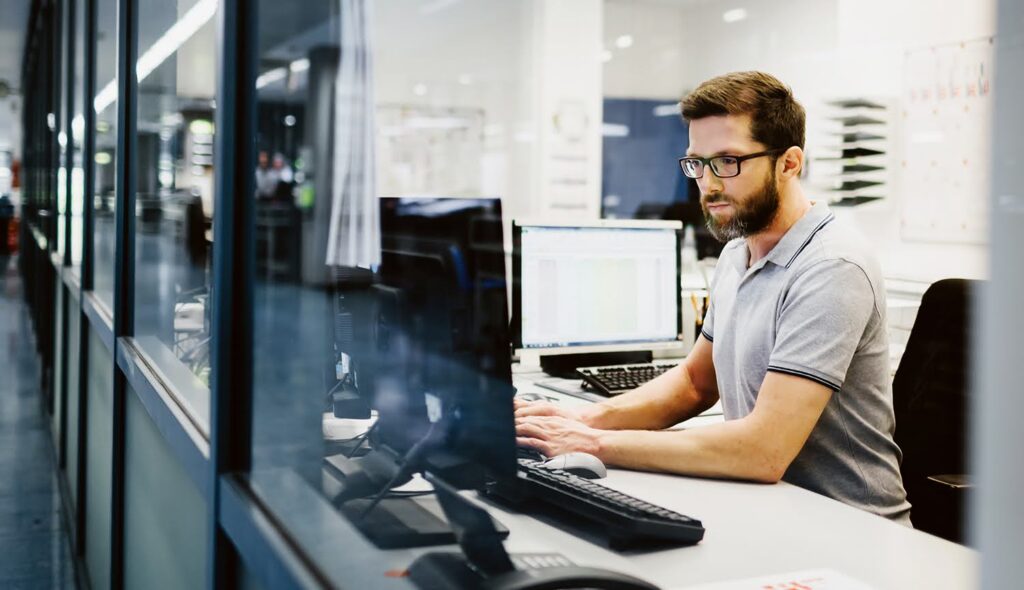 A young professional in a modern office concentrates on the computer screen in front of him while typing at a keyboard.