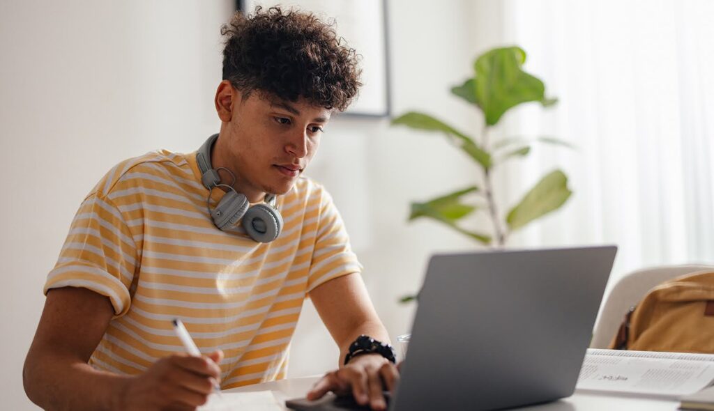 A young man with headphones around his neck looks at a laptop screen while taking notes on paper.