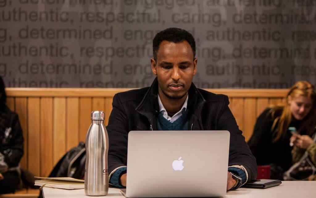 A student works on a laptop in a public space, surrounded by other students.