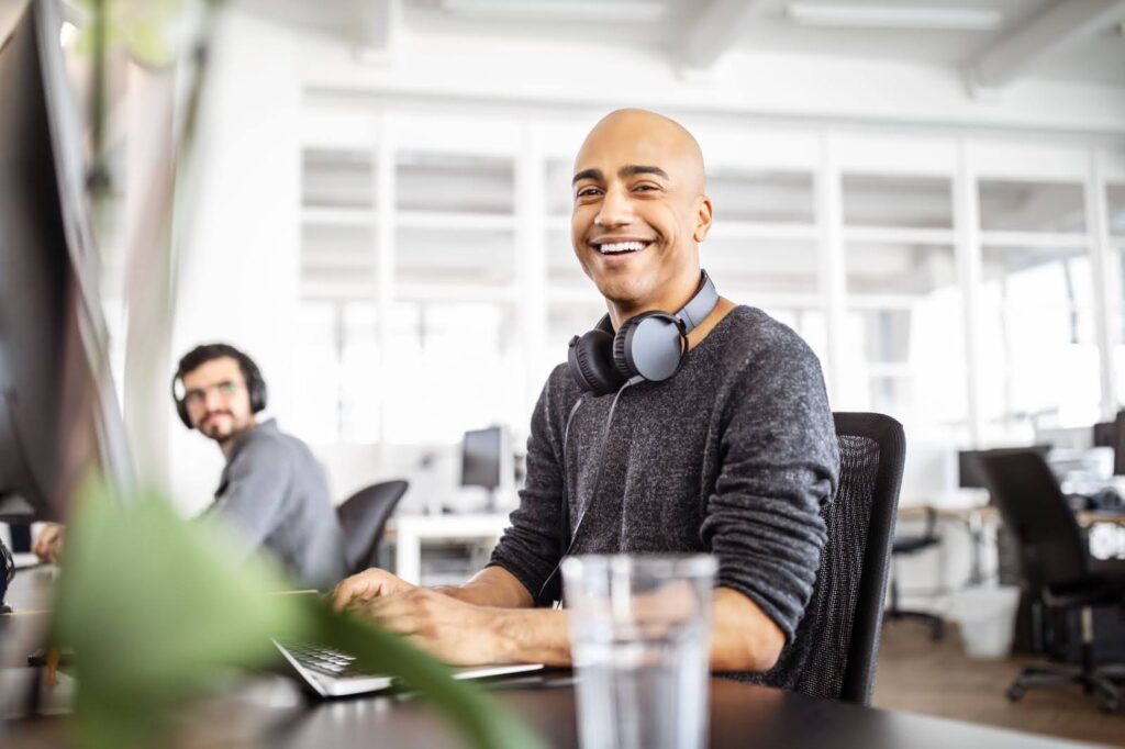 A computer science professional dressed in business casual attire smiles as they sit in front of a laptop and monitor in an open-plan office.