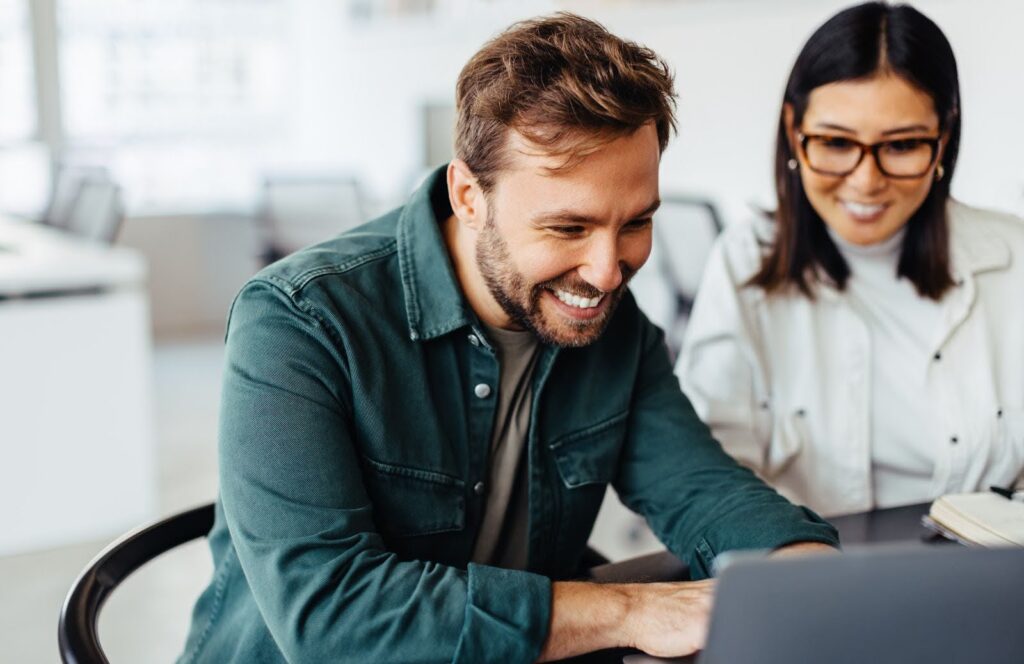 Two students smile at something they see on a laptop computer screen.