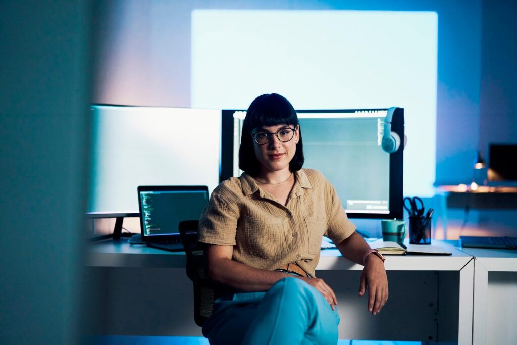 A woman at an office desk sits facing the camera; behind her on her desk are a large external screen and a laptop, both displaying coding text.