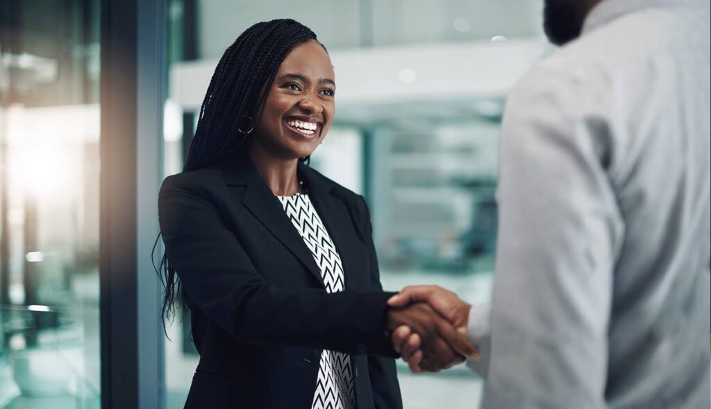 A smiling young professional woman shakes hands with a male business associate in an office.