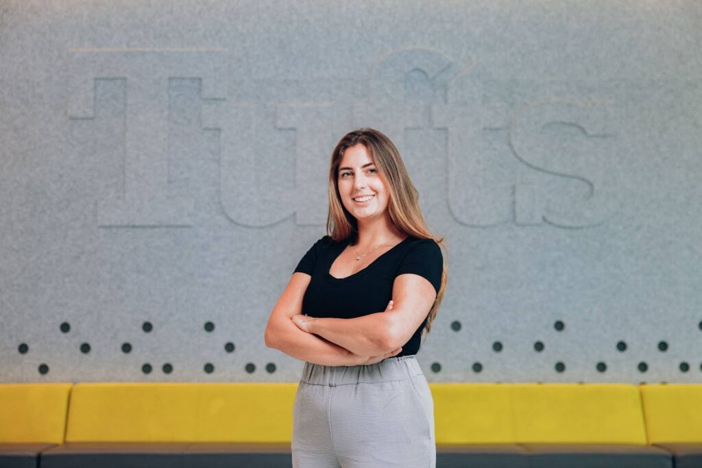 Hope Rogan, smiling and with arms crossed, stands in front of a large Tufts logo.