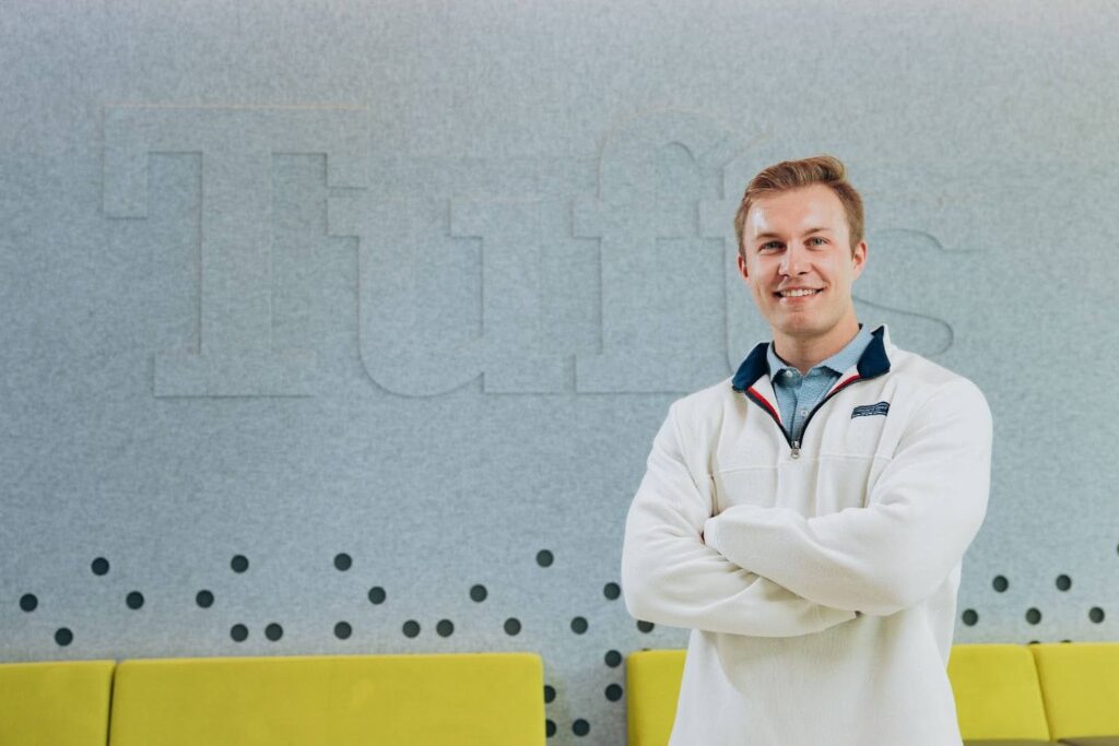 Derik Hendricksen, smiling and arms folded, stands in front of a large Tufts logo.