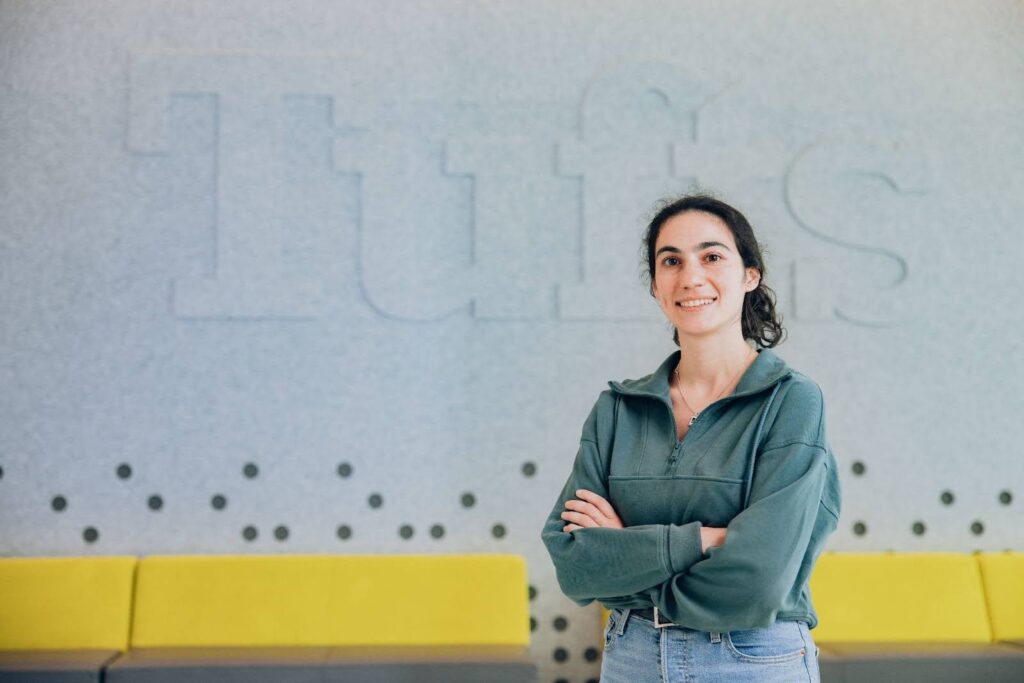 Lanie Kropp stands, arms folded and smiling, in front of a large Tufts logo.