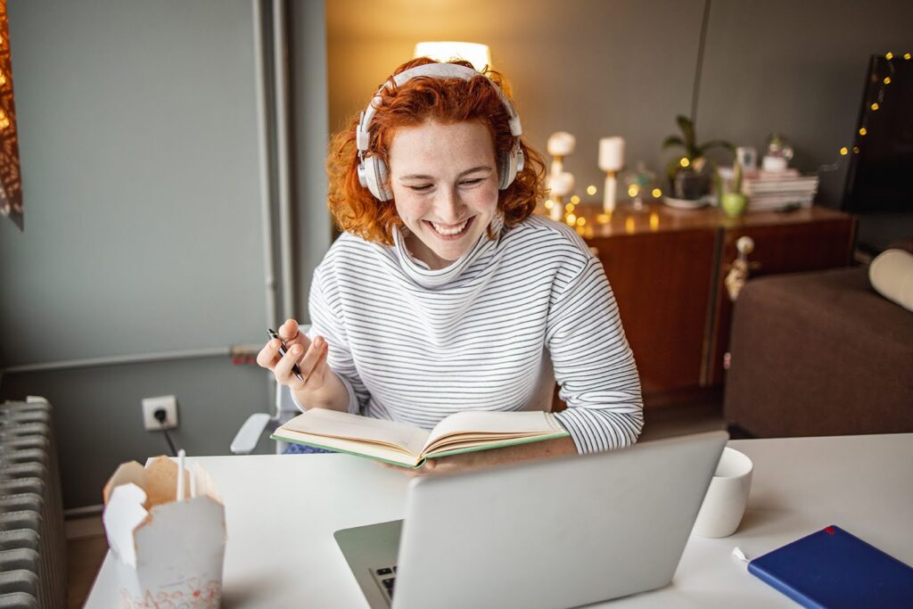 A smiling student wearing headphones holds an open book while looking at a laptop computer screen; she is seated at a dining room table.