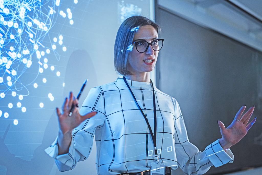 A young professional stands in front of a projection screen displaying back-end programmers’ code; she is explaining the information on the screen.