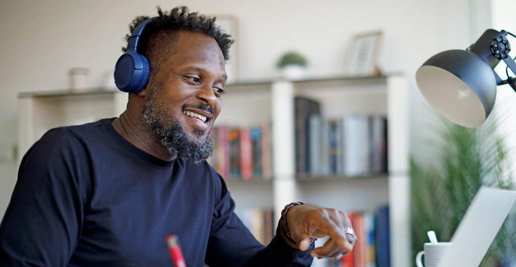 A man wearing headphones sits in a home office and points to his laptop screen with his left hand; he uses a pencil to write in a notebook with his right hand.